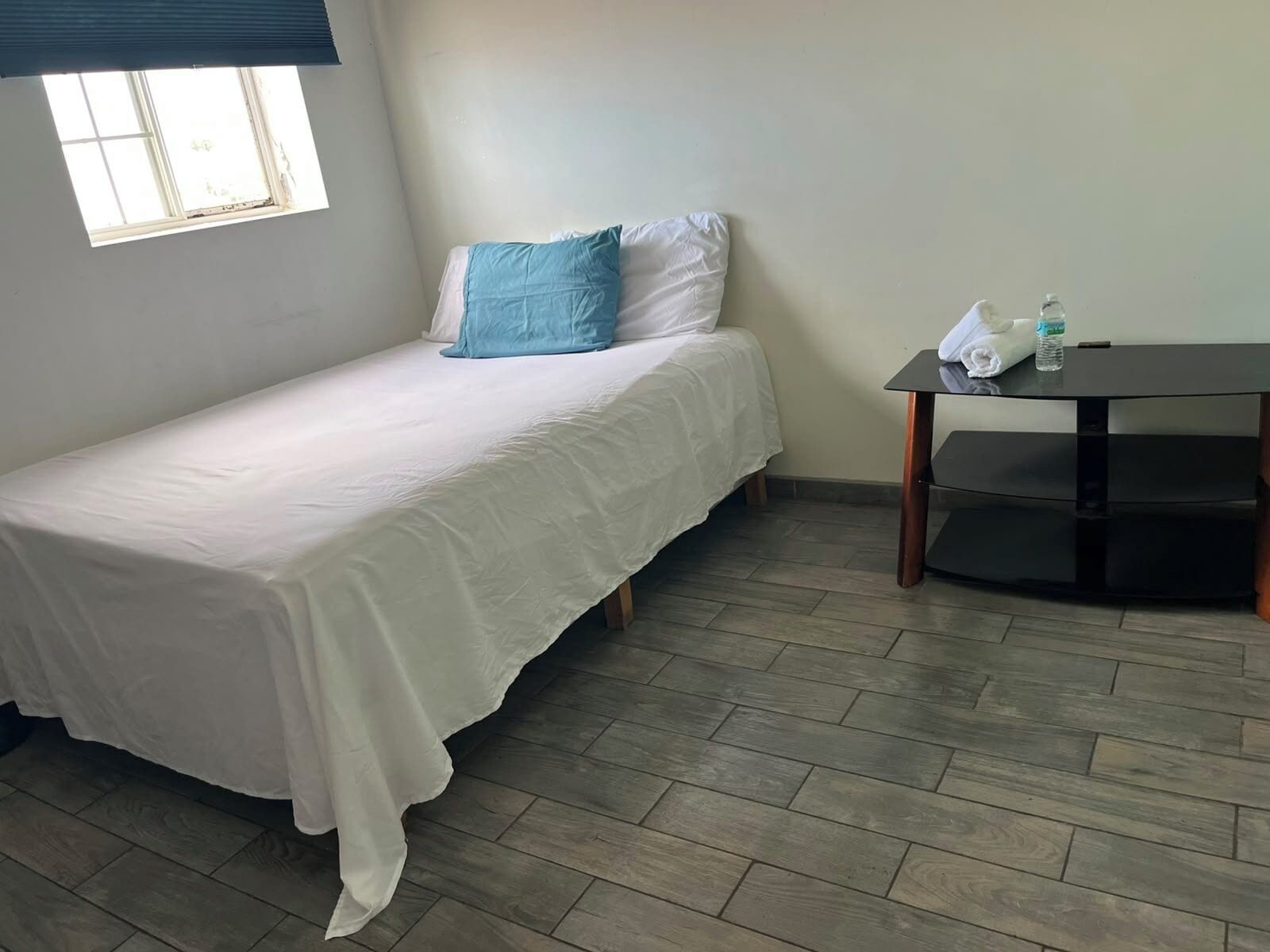 Uma cama de solteiro com colcha branca e travesseiro azul, uma mesinha com uma garrafa e toalha, e uma janela com persianas.