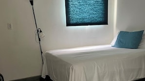 Ein Einzelbett mit weißer Matratze und blauem Kopfkissen, eine stehende Stehlampe und ein Fenster mit Verdunklung.