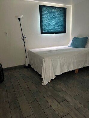 Una cama individual con colchón blanco y almohada azul, una lámpara de pie y una ventana con persiana oscura.