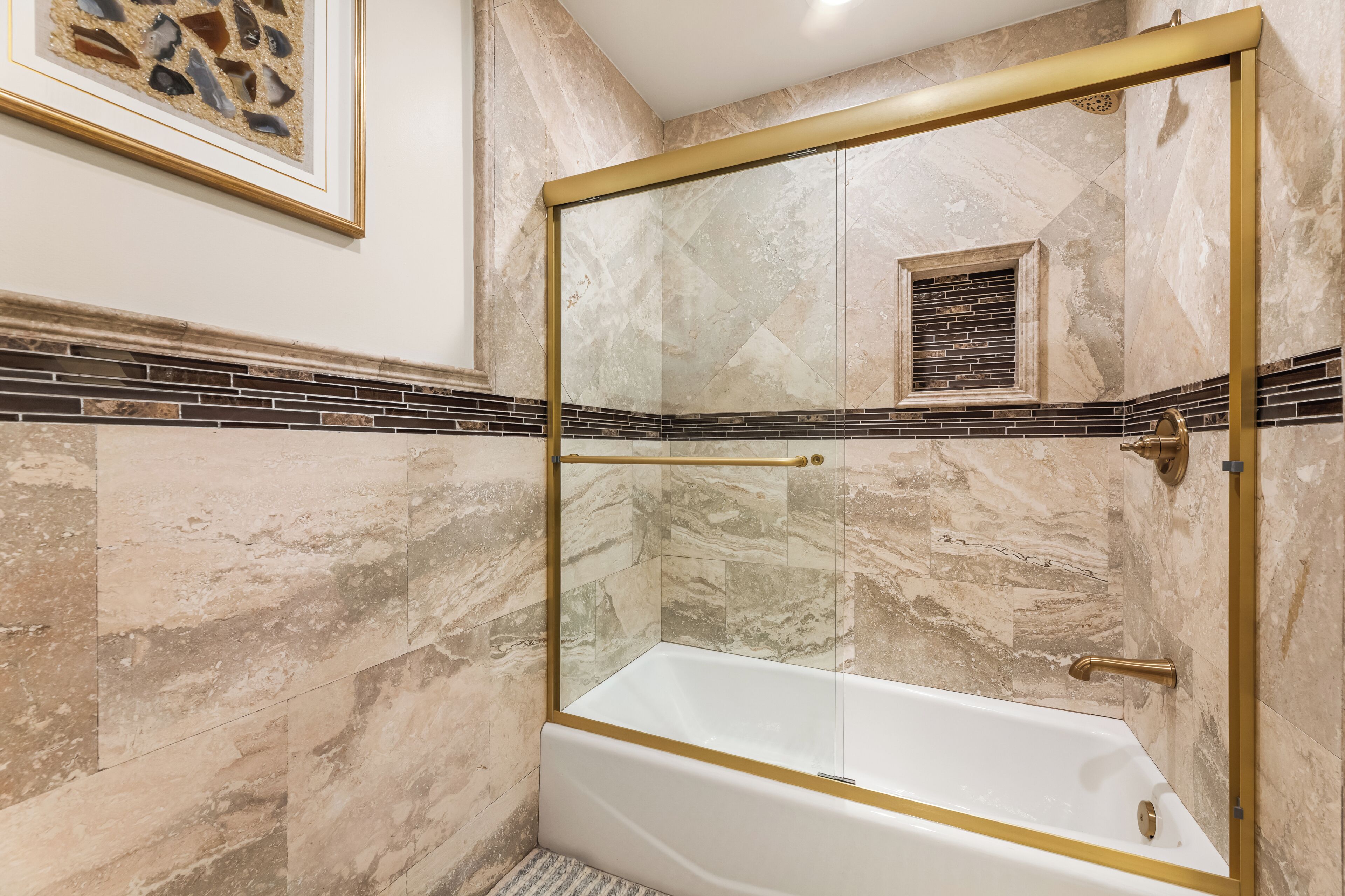 Un baño con ducha envuelta en vidrio, paredes de mármol y una obra de arte enmarcada sobre el espejo.