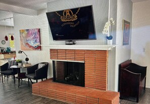 A modern living room with a flat-screen TV mounted above a brick fireplace, a dining area with chairs, and a large abstract painting on the wall. - Marvelous 3-bedroom house in sunny Palm Springs (Palm Springs)