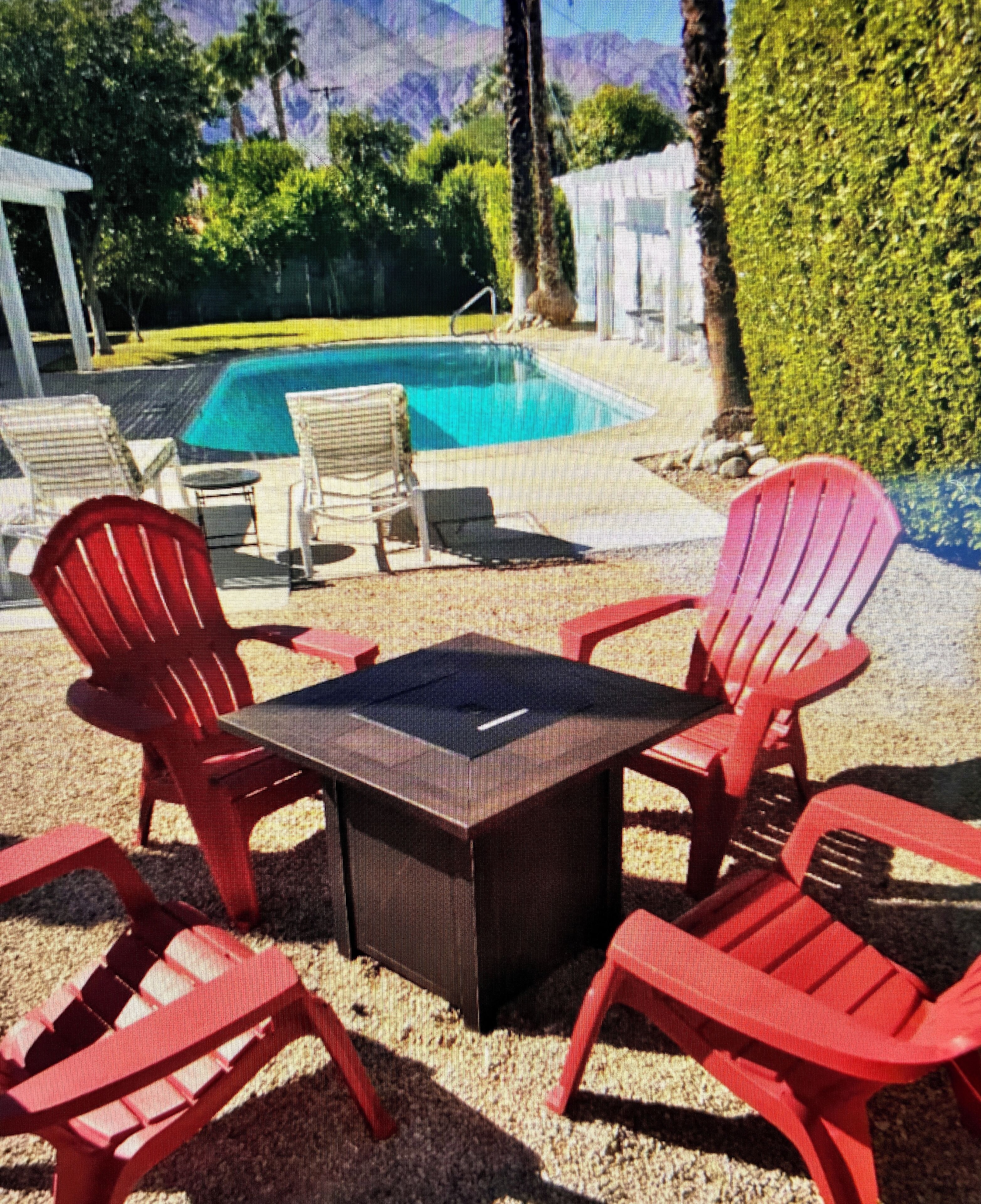 A pool area with a table and chairs, surrounded by a hedge and a mountain view.