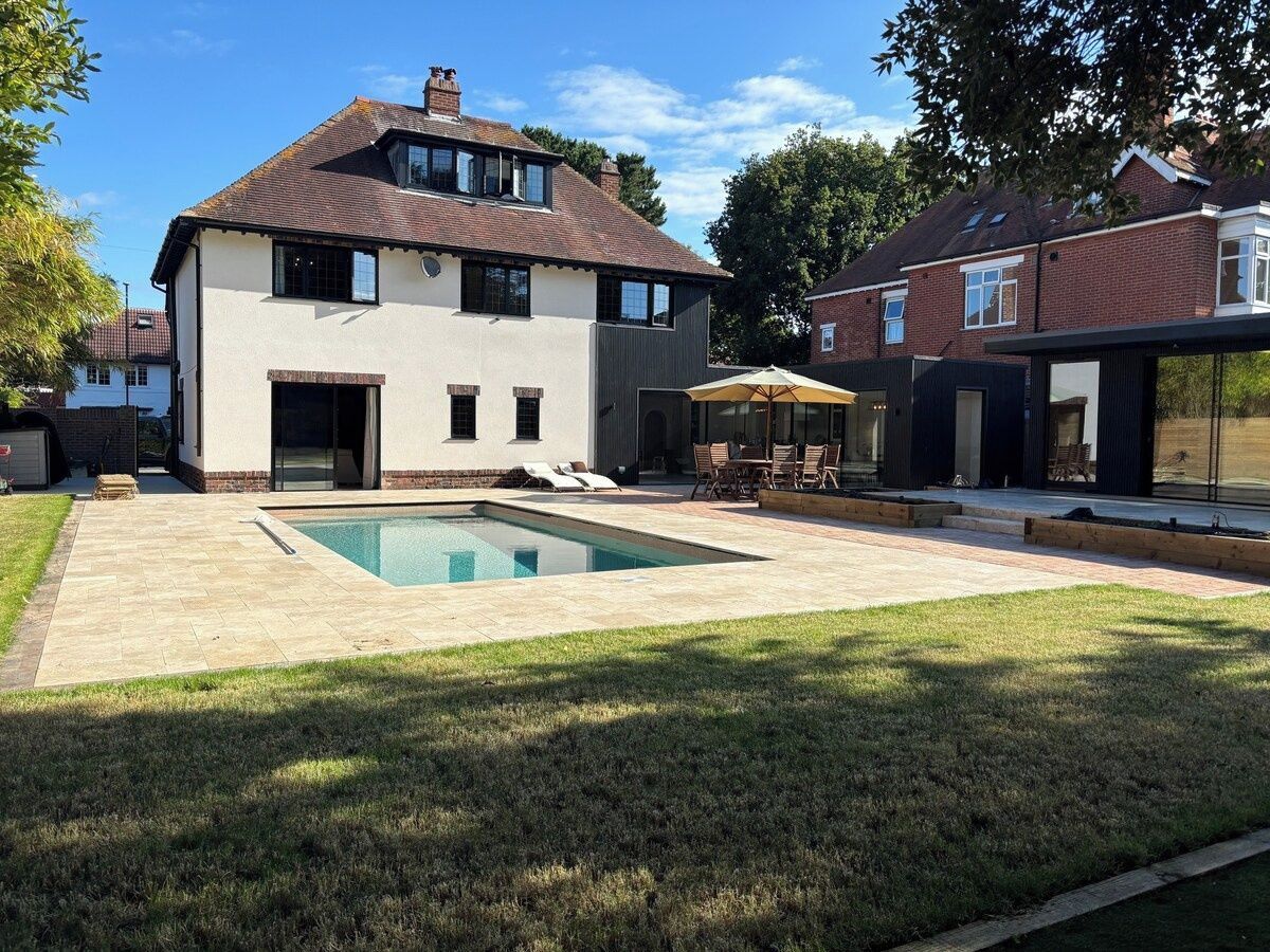 A two-story house with a swimming pool, a patio with chairs, and a lawn.