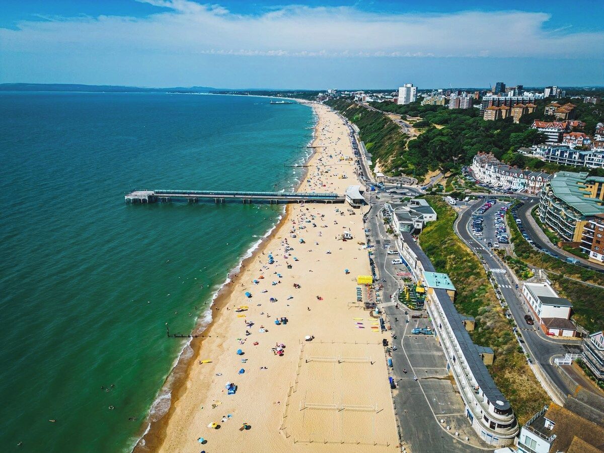 An aerial view of a beach with a pier extending into the sea, adjacent to a road with buildings and a cityscape in the background.
