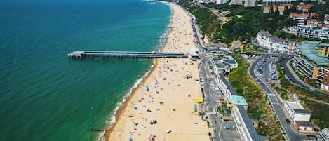 An aerial view of a beach with a pier extending into the sea, adjacent to a road with buildings and a cityscape in the background.