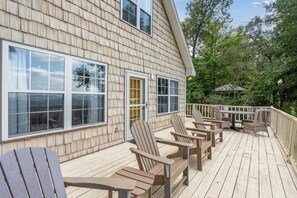 Eine Holzterrasse mit Adirondack-Stühlen und einem Tisch, mit Blick auf ein bewaldetes Gebiet.