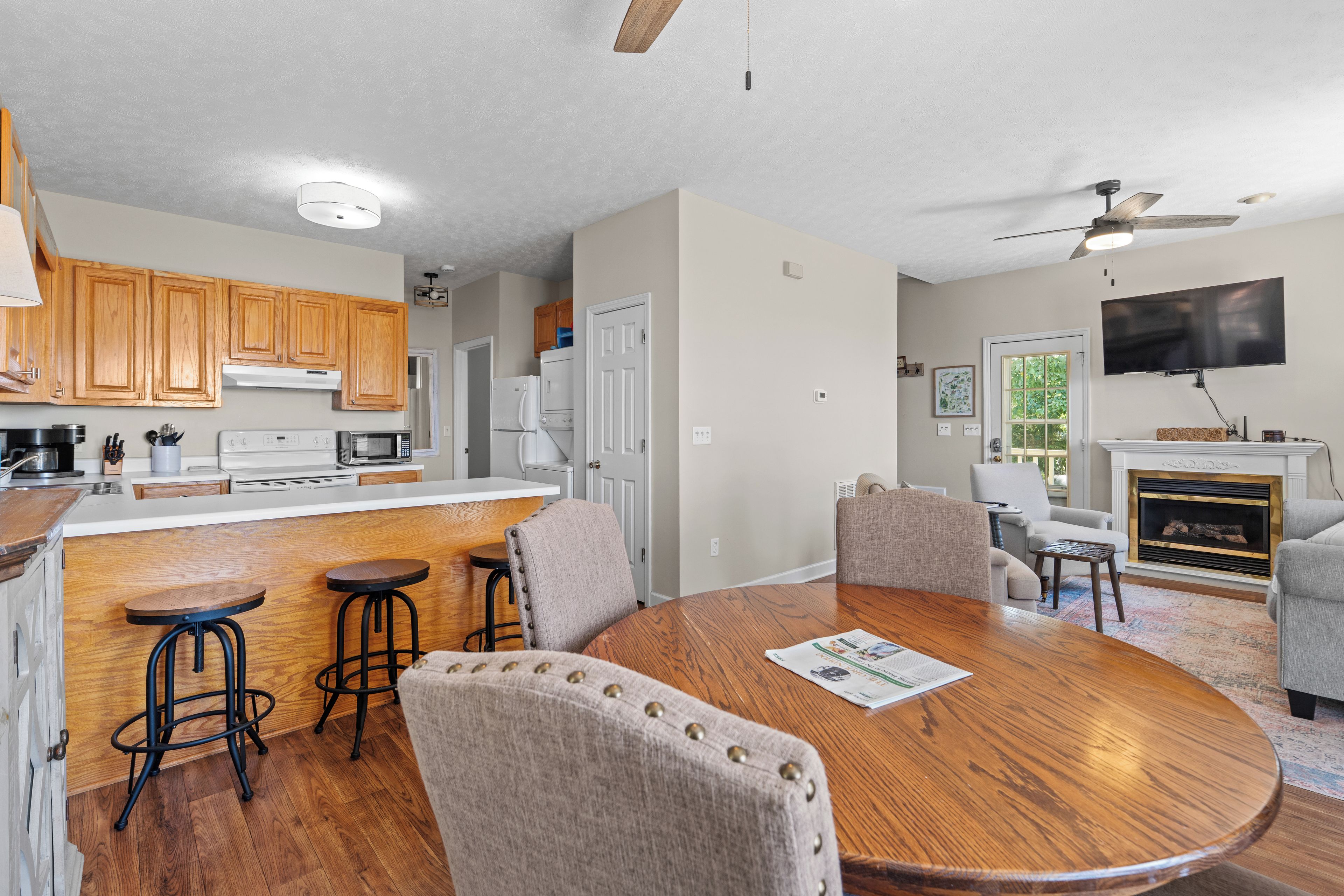 A modern kitchen with wooden cabinets, a dining area with a round table and bar stools, a living room with a fireplace, and a ceiling fan.