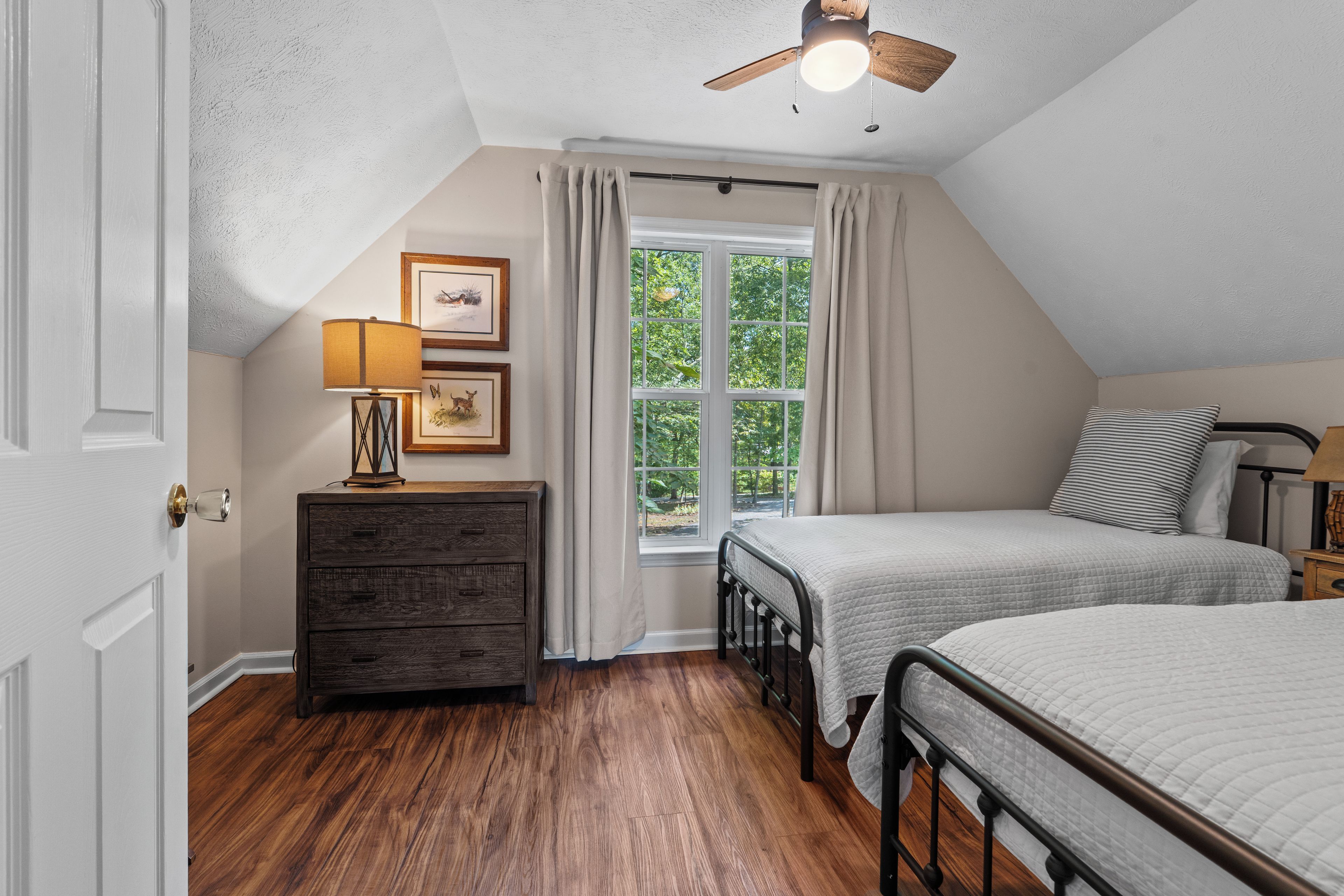 A bedroom with a sloped ceiling, a window with curtains, a bed with a headboard, a dresser, and a lamp.