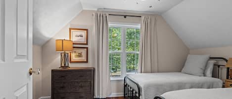 A bedroom with a sloped ceiling, a window with curtains, a bed with a headboard, a dresser, and a lamp.