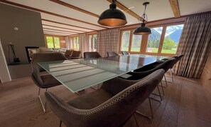 A modern dining area with a glass-top table surrounded by leather chairs, wooden beams on the ceiling, and large windows with curtains.