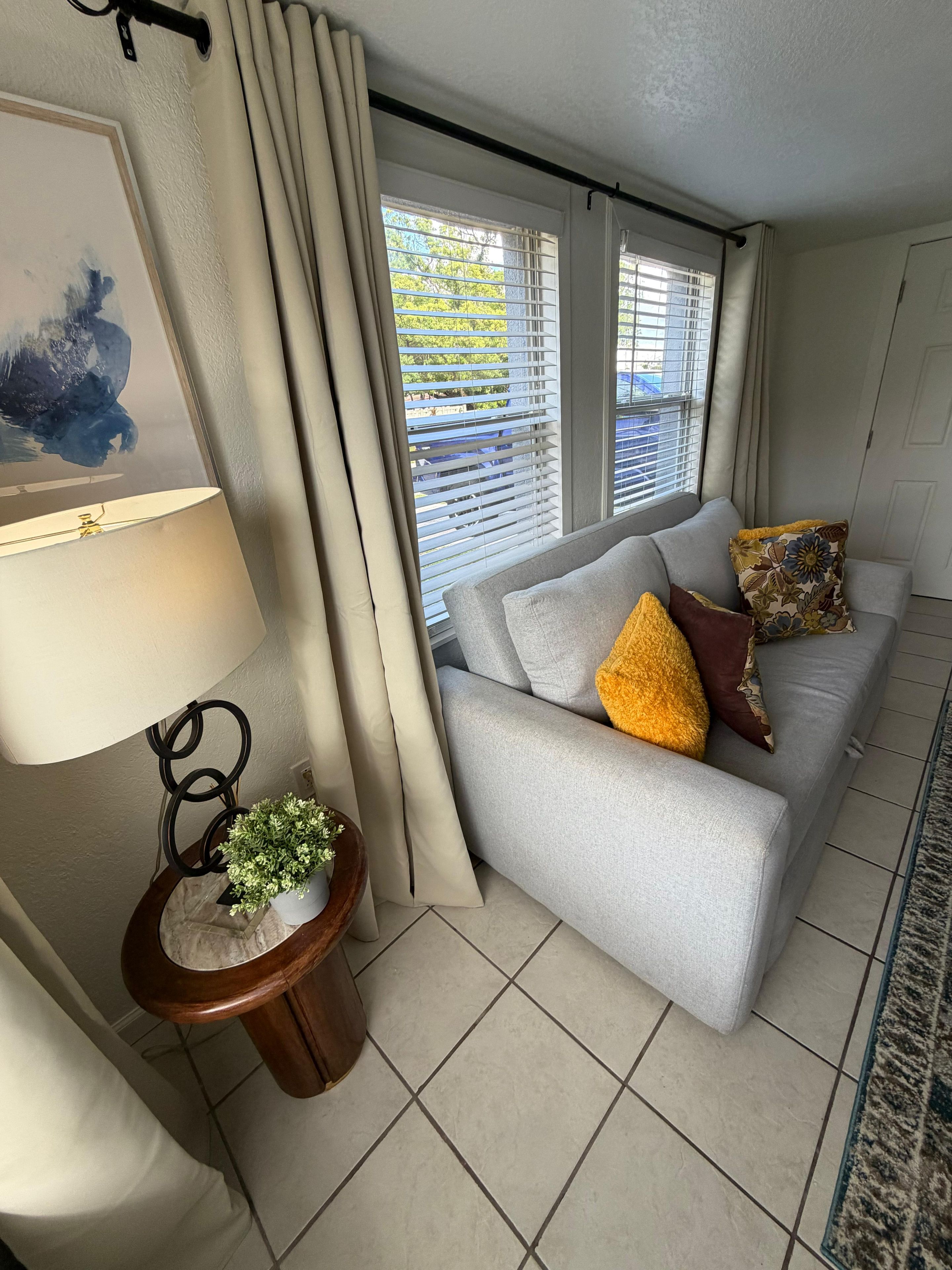 Sala de estar com um sofá cinza, uma pequena mesa lateral com uma planta, um quadro emoldurado e uma janela com cortinas.