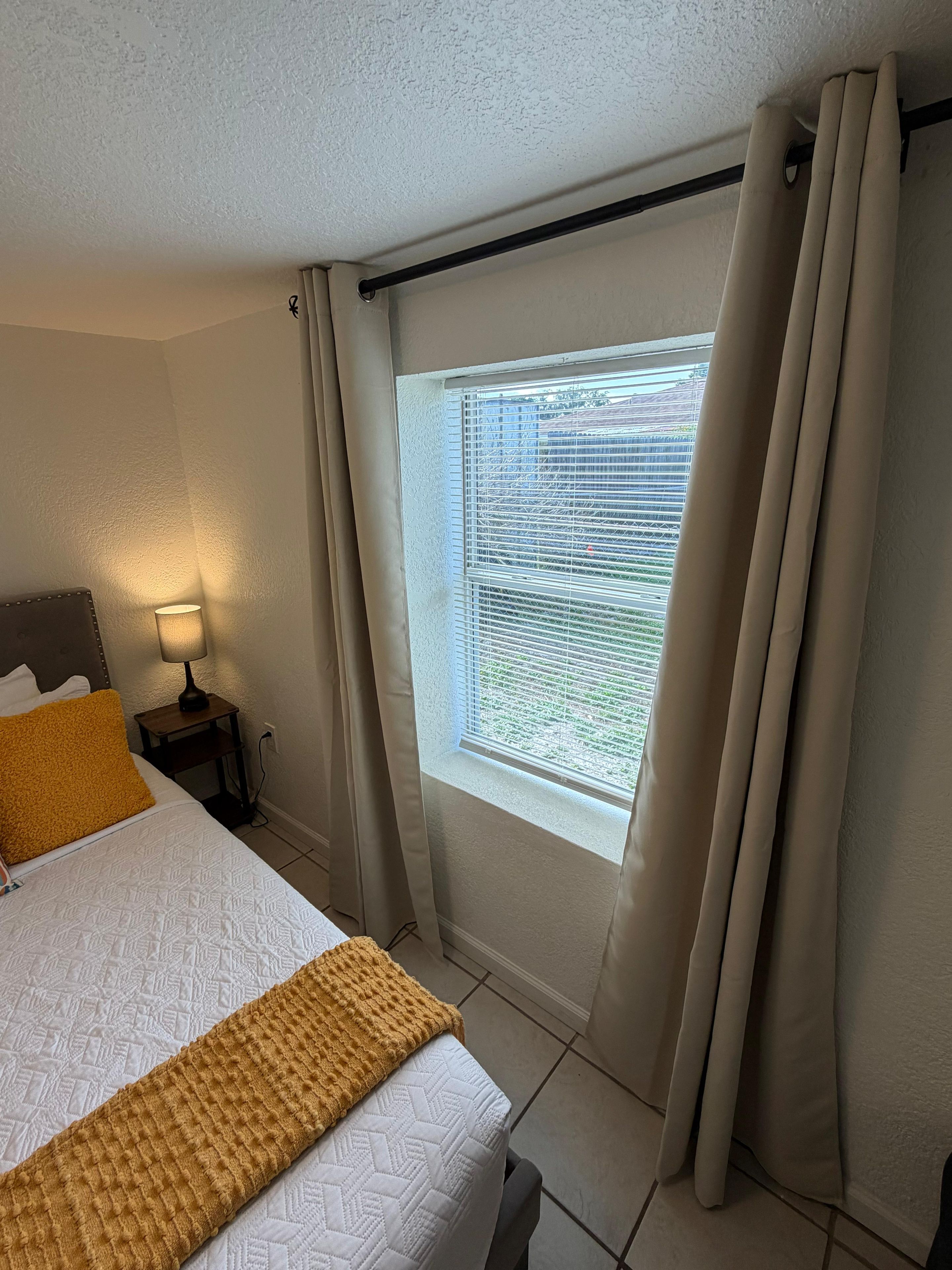 Quarto de hotel com cama, mesa de cabeceira com luminária, janela com cortinas e vista para o exterior.