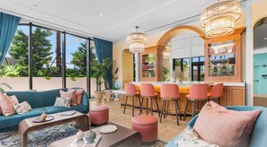 A modern living room with a sofa, coffee table, and ottoman, a bar area with stools, and a large window with a view of trees. - Nomad Residences Wynwood Art Trendy Area (Miami)