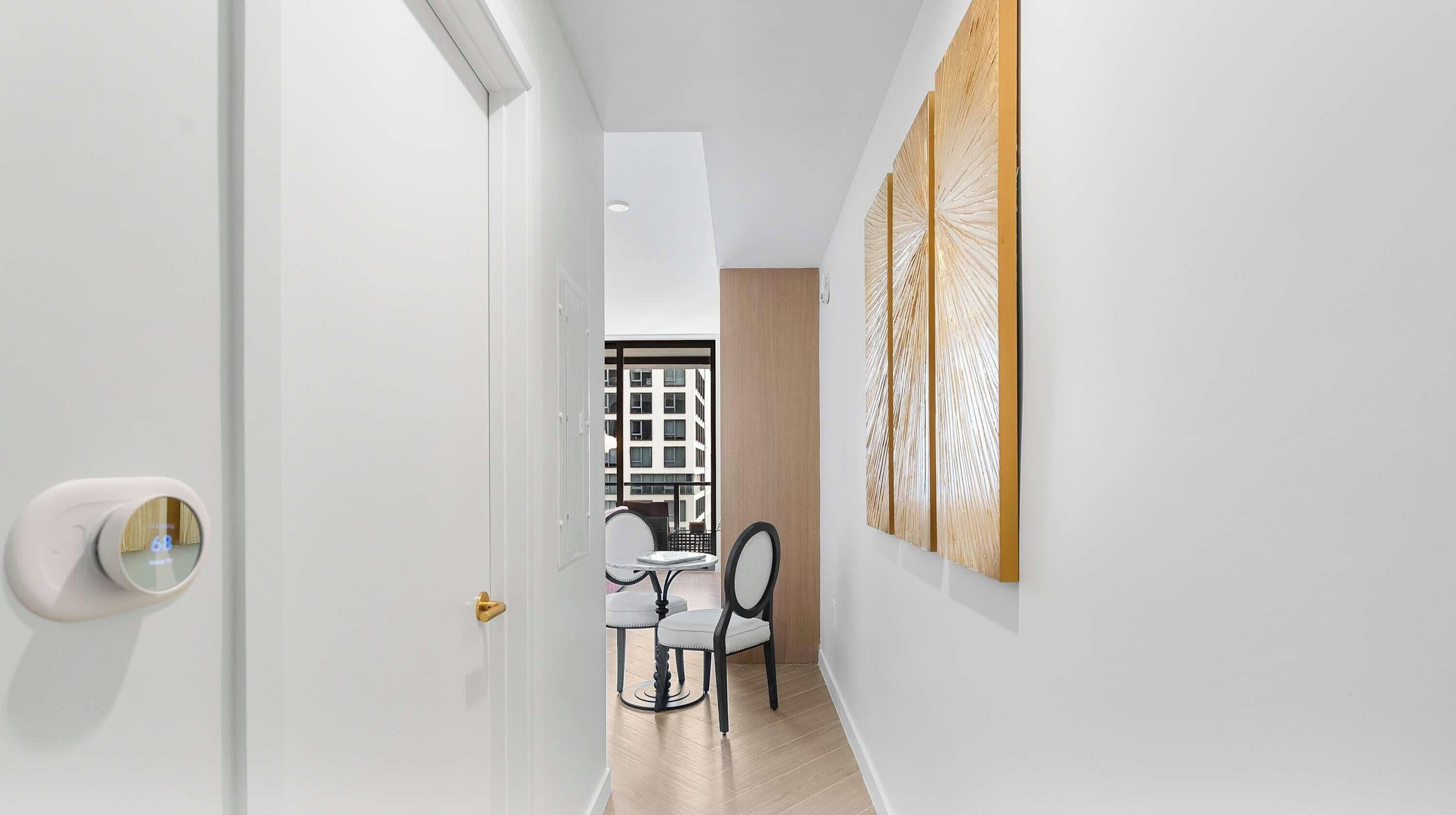 Un couloir avec une porte blanche, un coin repas avec une table et des chaises, et une vue sur un immeuble par la fenêtre.