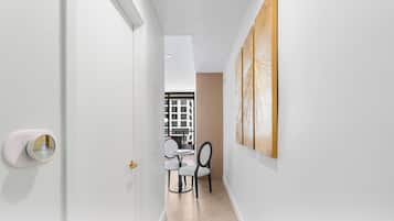 Un couloir avec une porte blanche, un coin repas avec une table et des chaises, et une vue sur un immeuble par la fenêtre.