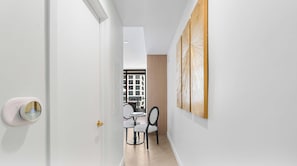 Um corredor com uma porta branca, uma área de refeições com mesa e cadeiras, e vista para um edifício através de uma janela.