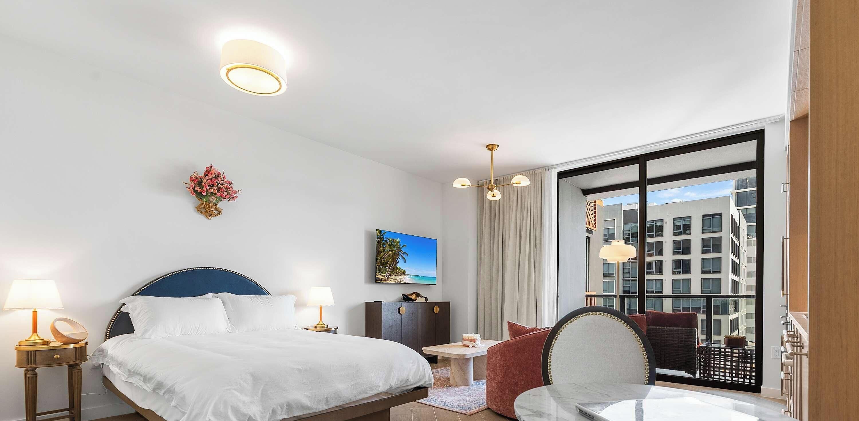 Une chambre d’hôtel avec un lit, des lampes de chevet, un bureau, une chaise, une vue sur les bâtiments et un tableau au mur.