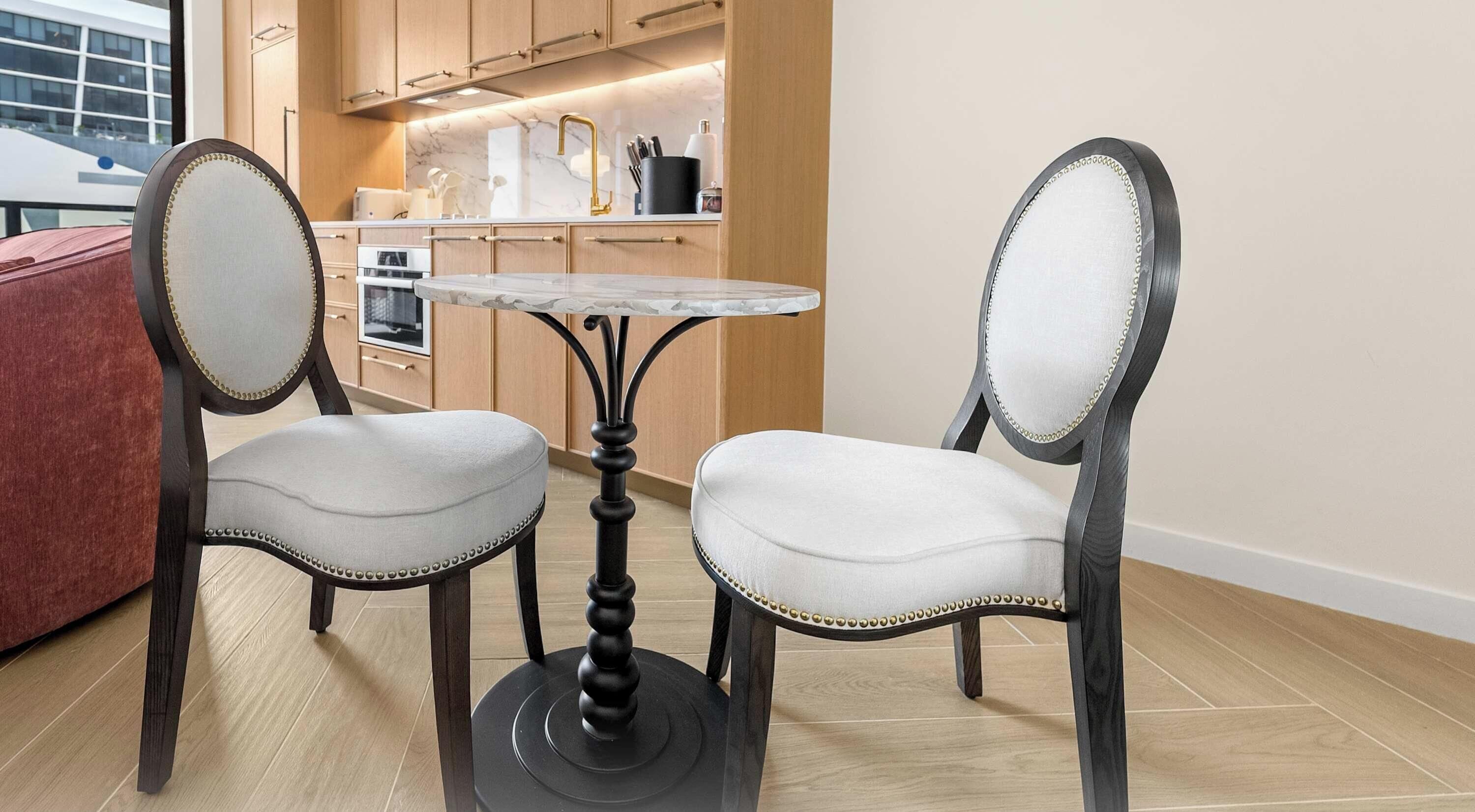 A dining area with two upholstered chairs, a round table, and a kitchen in the background.