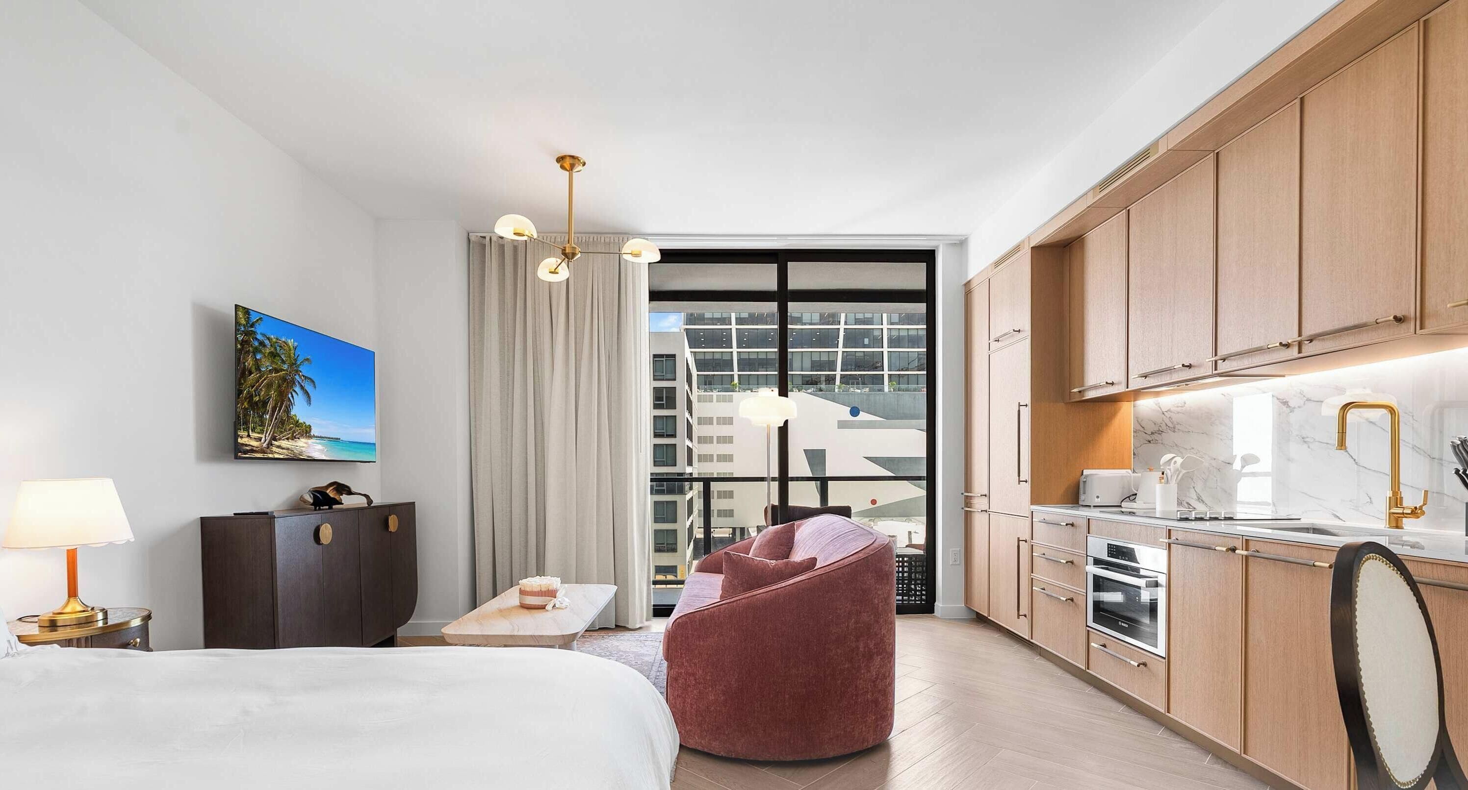 A hotel room with a bed, a desk, a chair, a kitchenette, and a large window with a city view.