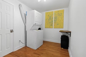 A laundry room with a white washer and dryer, a wooden floor, a door, and two abstract yellow paintings on the wall.