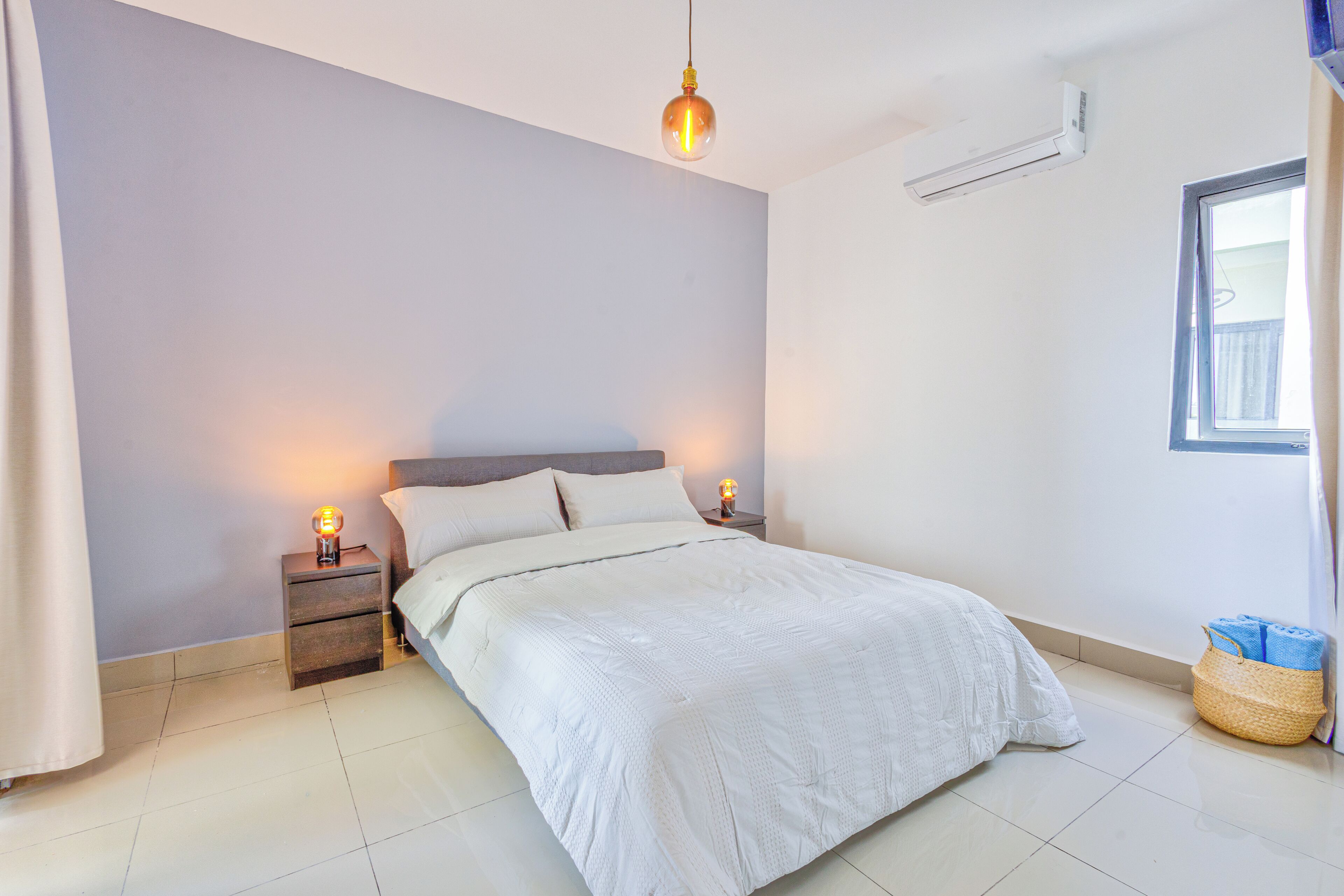 A bedroom with a bed, two bedside lamps, a hanging light, a window, and a wall-mounted air conditioner.