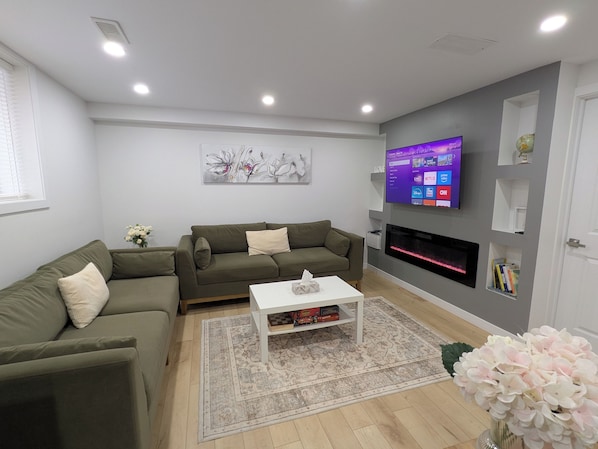 A modern living room with a sectional sofa, a flat-screen TV mounted on the wall, a fireplace, and a coffee table with books. - 3 Bedroom Modern, Stylish & Spacious Home with King Size Bed! (Stratford)