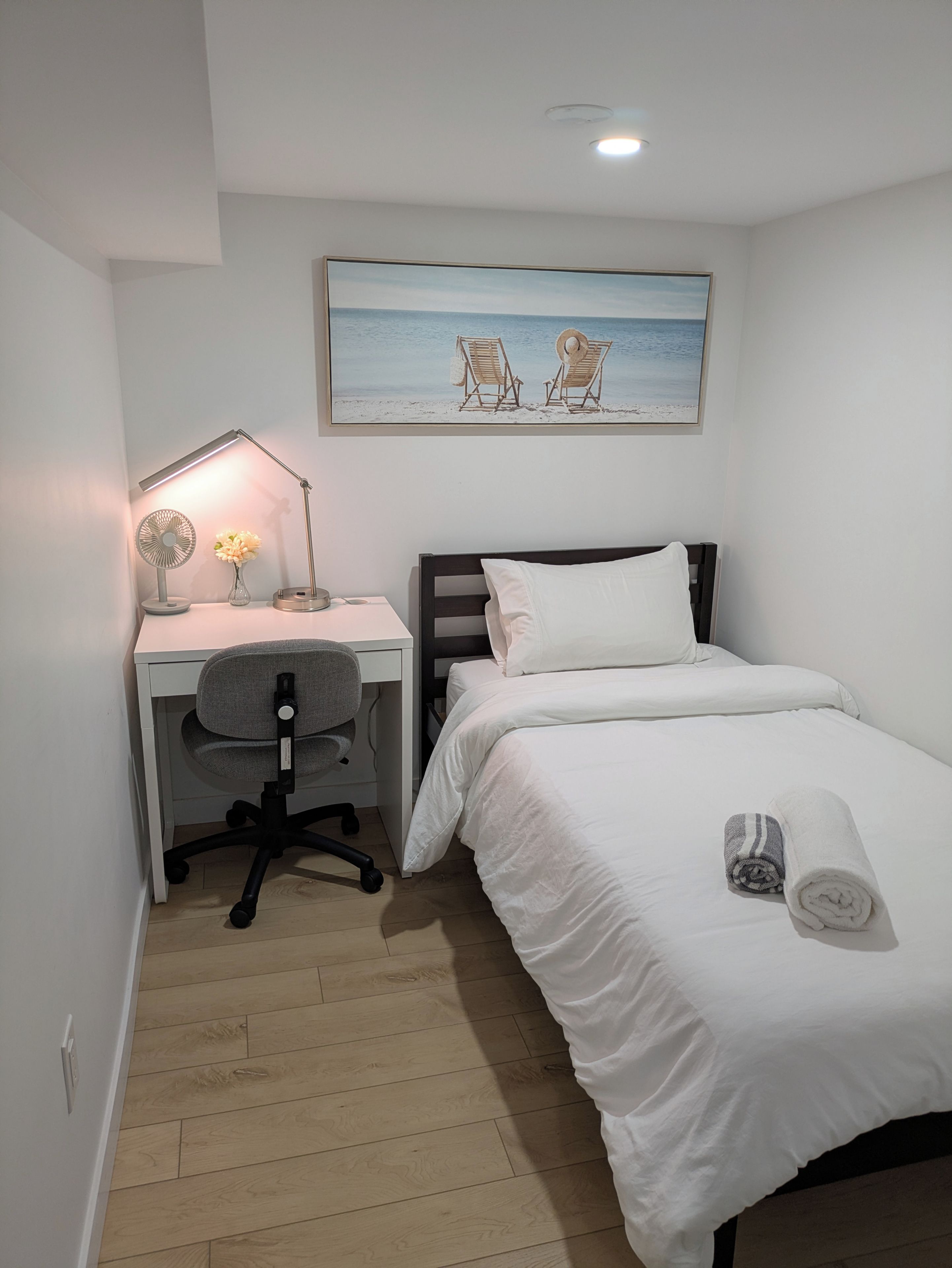 A bedroom with a single bed, a desk with a chair, a standing lamp, and a painting of beach chairs.