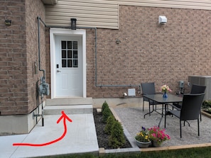 A brick building with a white door, outdoor seating with a table and chairs, and a small garden area with flowers. - 3 Bedroom Modern, Stylish & Spacious Home with King Size Bed! (Stratford)