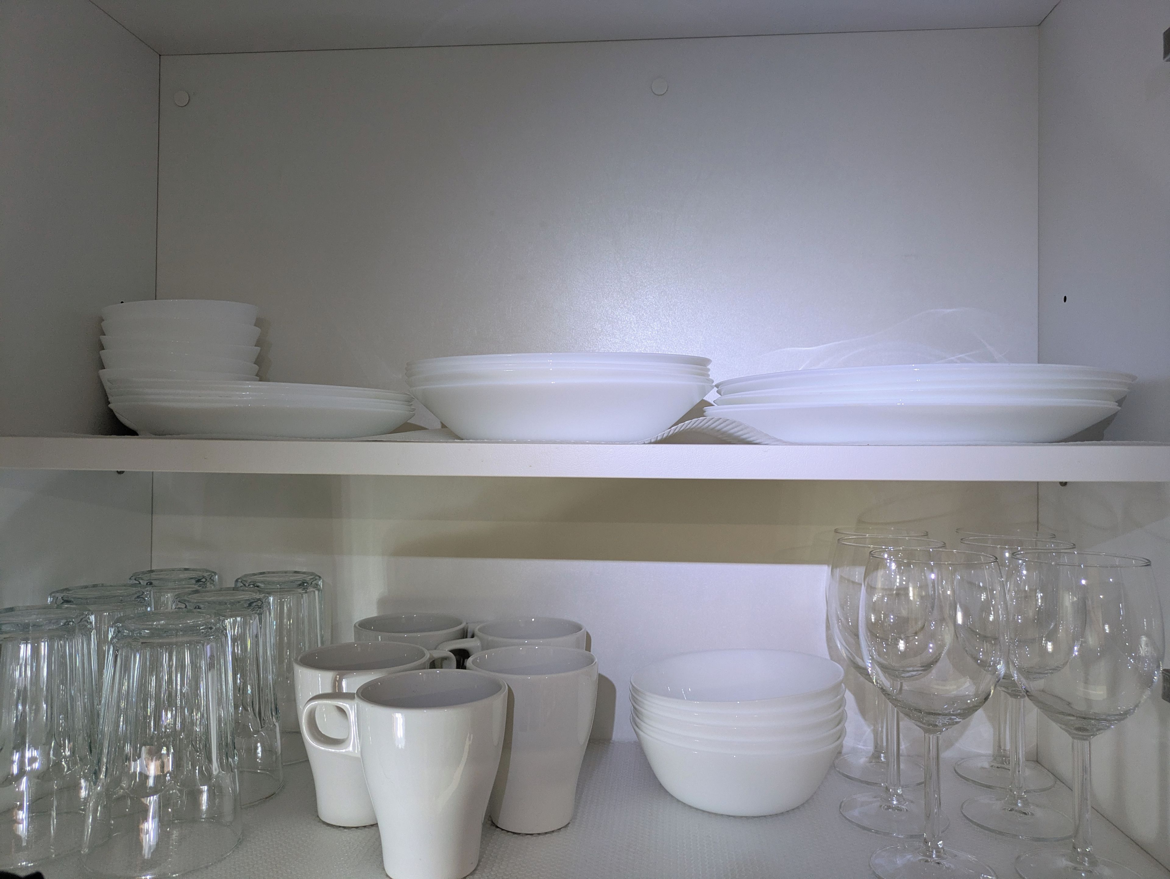 Una estantería blanca con platos y tazones blancos apilados, y debajo, vasos de vidrio y copas de vino.