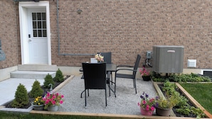 Eine Terrasse mit zwei Stühlen, einem Tisch, einer Ziegelwand, einer Tür und einer Klimaanlage.