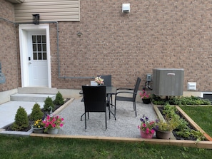 A patio with two chairs and a table, a brick wall, a door, and an air conditioning unit. - 3 Bedroom Modern, Stylish & Spacious Home with King Size Bed! (Stratford)