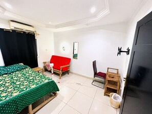 A room with a bed, a red sofa, a chair, a small table, a wall-mounted TV, and a door. - Room-beautiful Room With Private Toilet (Bamako)