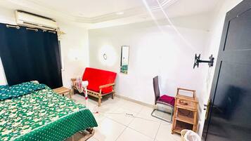 A room with a bed, a red sofa, a chair, a small table, and a trash bin.
