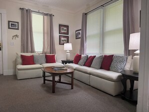 A living room with a sectional sofa, a coffee table, and a lamp. There are curtains on the windows, and framed pictures on the wall. - Downtown Aiken Enjoyable 3-bedroom house-walking distance to downtown (Aiken)