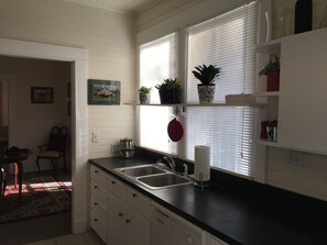 Una cocina con armarios blancos, encimera negra, pileta y ventana con persianas.