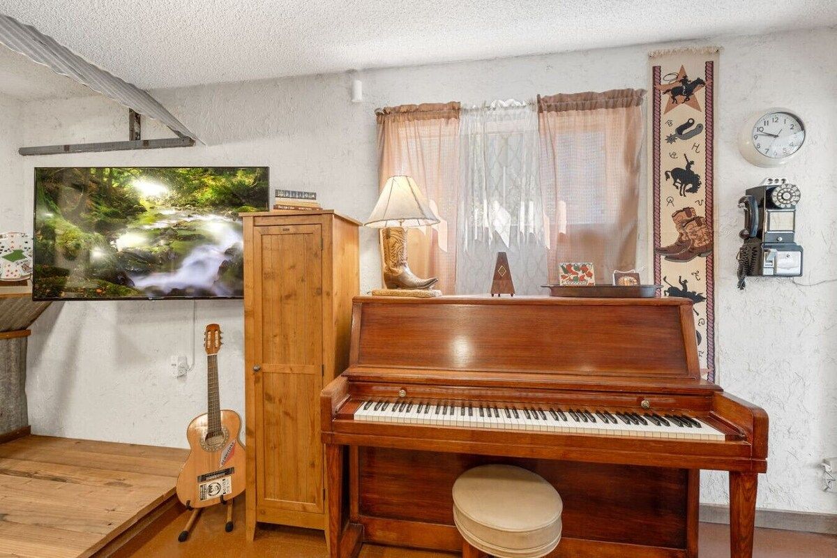 A living room with a wooden piano, a television, a guitar, and a wall clock.