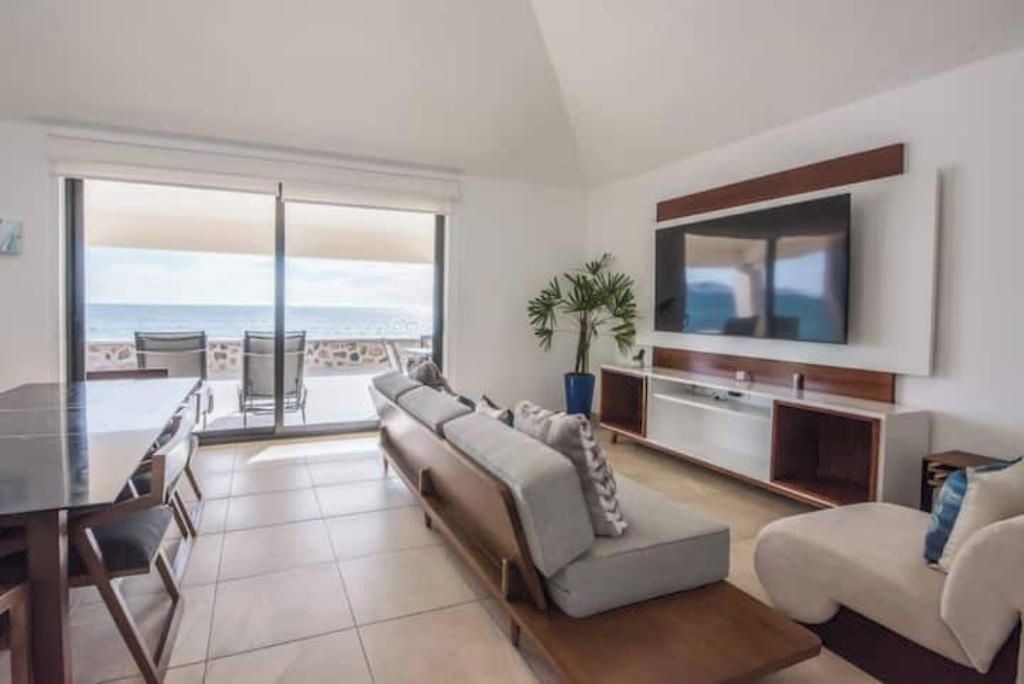 Una sala de estar moderna con un televisor grande, un sofá, una mesa de comedor y vistas al océano.