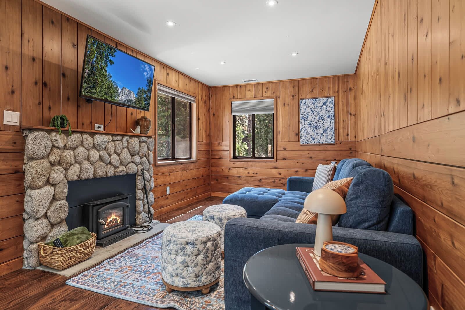A cozy cabin interior with a stone fireplace, a blue sofa set, and a television mounted on the wall.