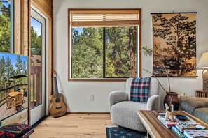 Ein Zimmer mit großem Fenster, einer Gitarre, einem Stuhl, einem Tisch und einem Gemälde.