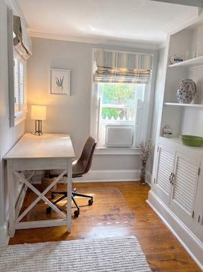 Una habitación con escritorio, silla, ventana con cortinas, radiador y estante con objetos decorativos.