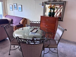 Quarto com mesa de jantar de vidro rodeada por cadeiras de metal, cômoda de madeira e cama com colcha roxa.
