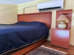 Cama com colcha azul escura, cabeceira de madeira, mesa de cabeceira com luminária e um aparelho de ar condicionado sobre a cama.