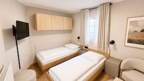 Ein Hotelzimmer mit zwei Einzelbetten, einem Fernseher, einem Fenster mit Vorhängen, einem kleinen Tisch und einem Sessel.