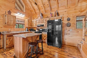 L’intérieur d’un chalet en bois comprend une cuisine équipée d’un réfrigérateur noir, de meubles de rangement en bois, d’un coin bar avec des tabourets et d’une grande fenêtre.