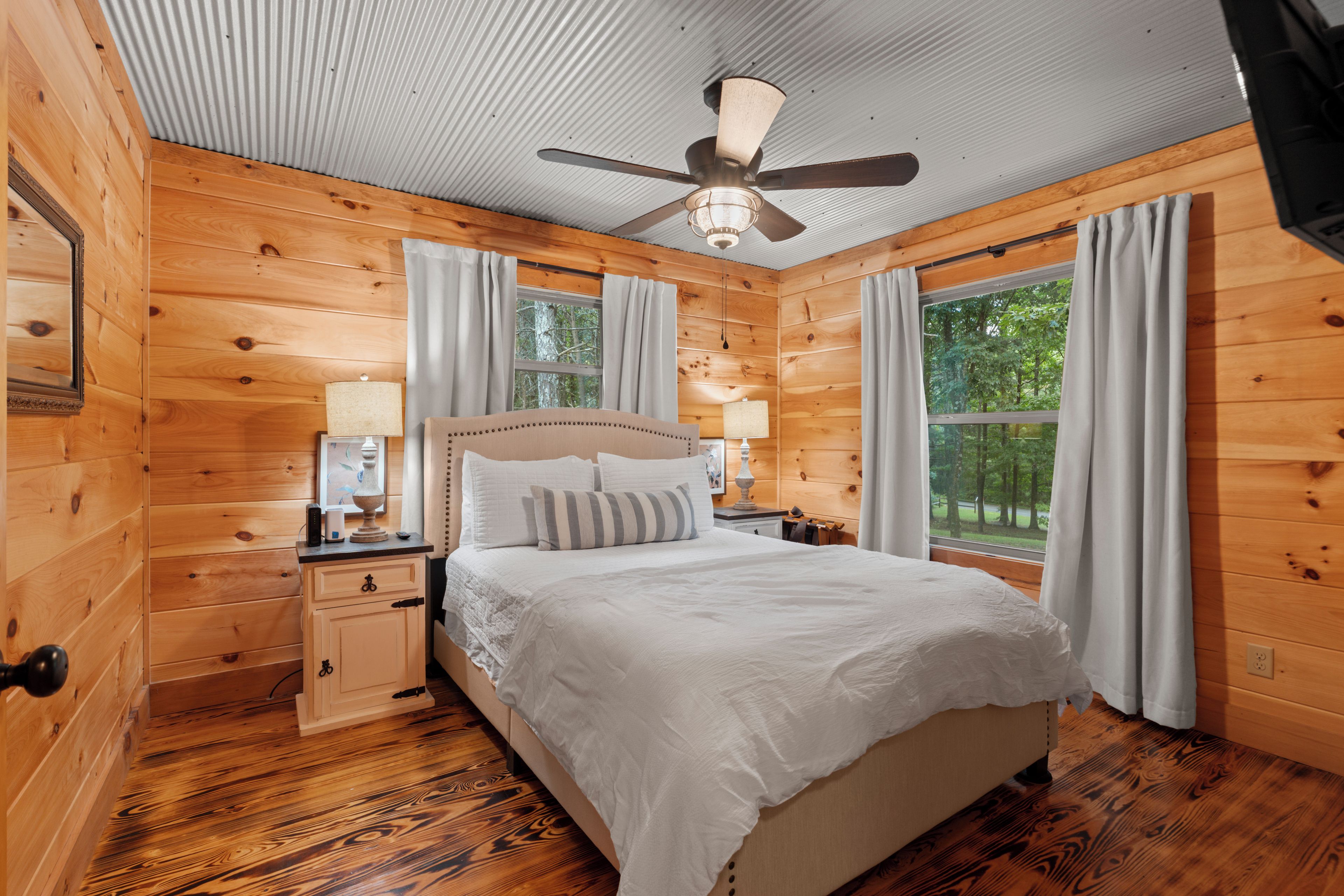 Ein Schlafzimmer in einer Holzhütte mit einem Bett, Nachttischen, einem Deckenventilator und einem Fenster mit Vorhängen.