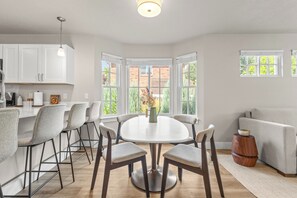 Une cuisine moderne avec des meubles blancs, un coin repas comprenant une table ronde et des chaises, et de grandes fenêtres donnant sur un bâtiment en briques.