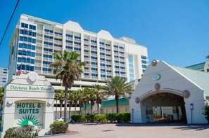 Entrada de hotel com placa indicando Daytona Beach Resort, área de estacionamento e um edifício alto com vários andares ao fundo.