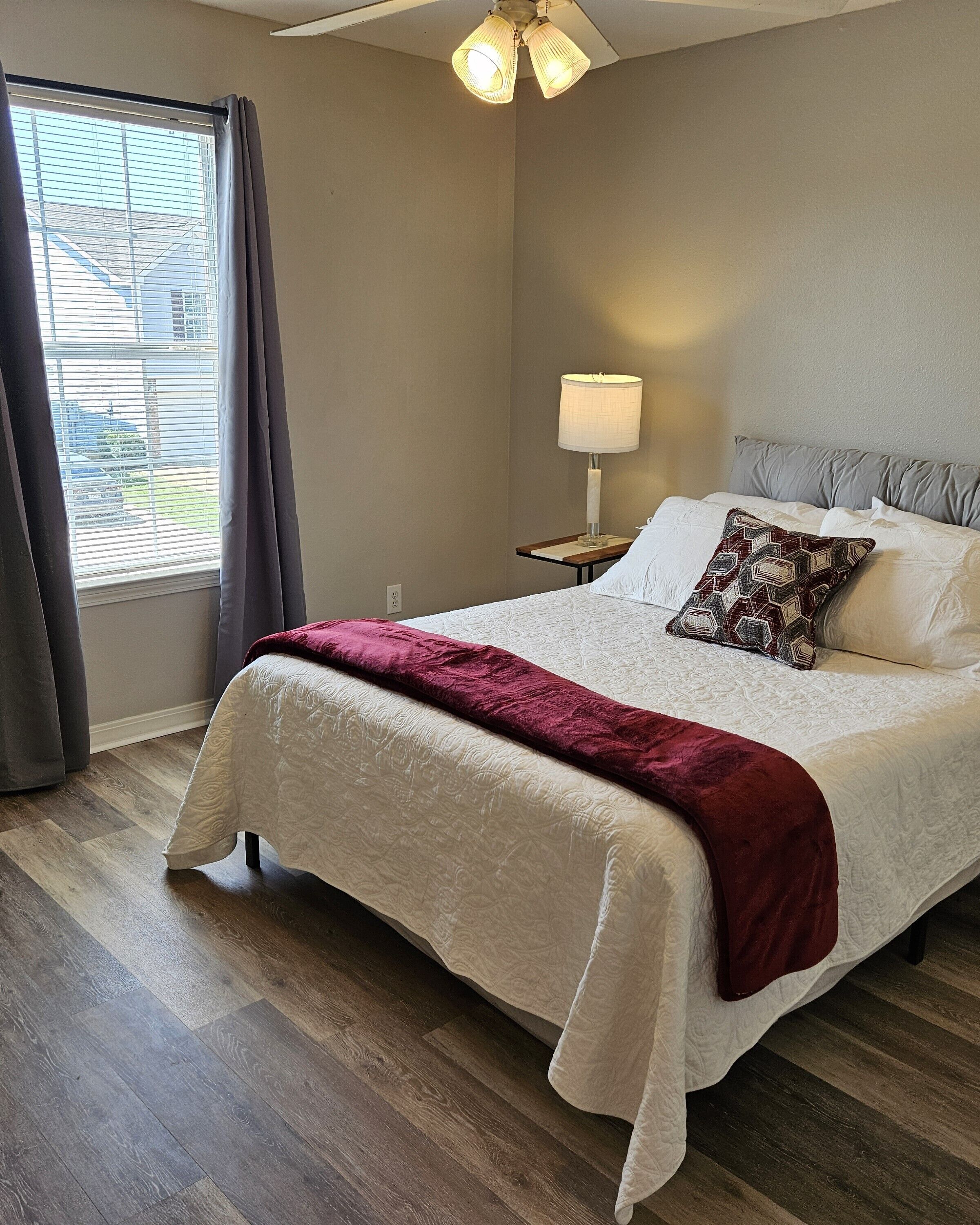Une chambre à coucher avec un lit, une table de chevet, une lampe et une fenêtre avec des rideaux.
