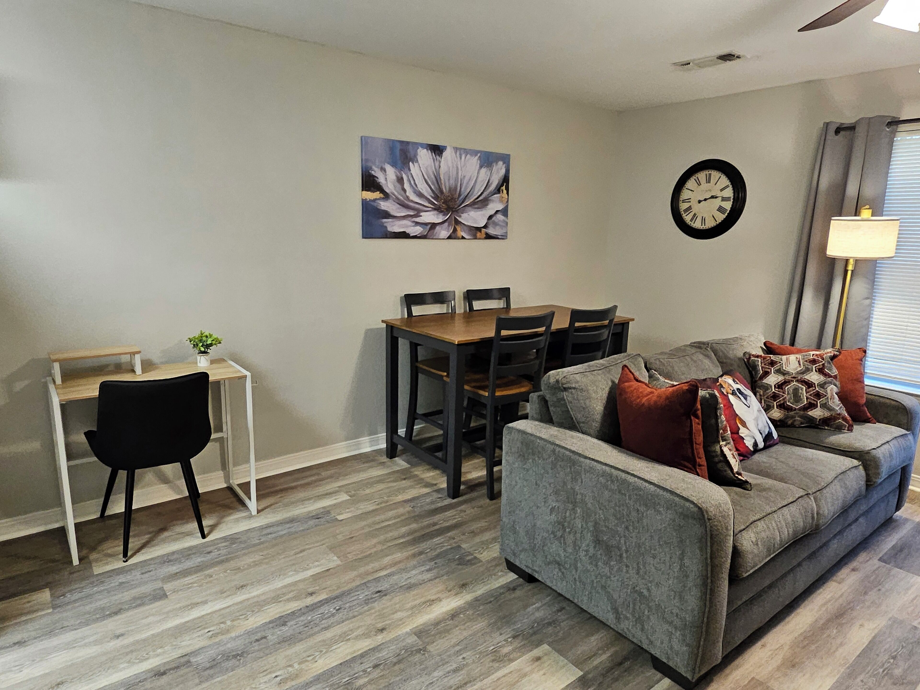 Un salon avec un canapé gris, une table à manger avec des chaises, un fauteuil noir, une horloge murale et un tableau représentant des fleurs.