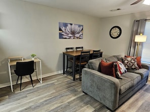 A living room with a grey sofa, a dining table with chairs, a black chair, a wall clock, and a flower painting. - Gameday Guesthouse 2BR/2BA updated townhome in fabulous Starkville 5 min to MSU (Starkville)